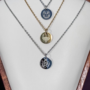 May include: Three layered necklaces featuring circular metal pendants. The top silver pendant displays a bat, the middle gold pendant shows a moth with a crescent moon, and the bottom silver pendant features a knight helmet with laurel branches.