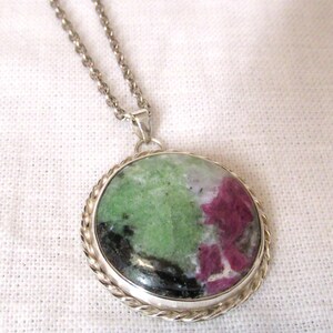 May include: A silver pendant necklace featuring a large, round gemstone. The stone displays a mix of green, pink, black, and white colors. The pendant is set in a silver frame with a twisted rope design and hangs from a delicate silver chain.