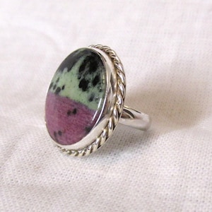 May include: Oval-shaped ring with a polished stone. The stone features a color block design with black, green, and magenta hues. The ring has a silver band and a twisted rope-like border around the stone.
