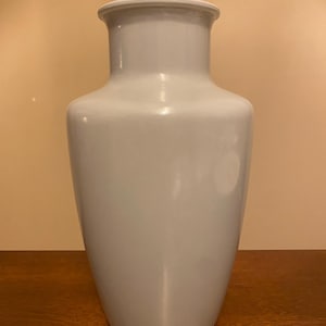 May include: A smooth, light gray ceramic vase with a glossy finish. The vessel features a wide, rounded body that tapers toward a narrow neck, ending in a flat, circular rim. The surface reflects light, highlighting its uniform color and shape.