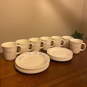May include: A set of off-white ceramic mugs and matching saucers featuring a delicate blue floral vine pattern around the rims. The collection includes several mugs with side handles and stacks of circular plates with a thin blue border design.