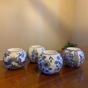 May include: Four spherical ceramic candle holders featuring white bases with intricate blue floral patterns and subtle yellow accents. Each holder has a circular opening at the top and a smooth, rounded exterior surface decorated with various botanical designs.