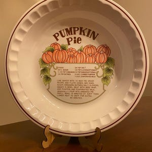 May include: A white ceramic pie dish with a fluted rim and a brown border. The center features a graphic of orange pumpkins with green leaves and the text Pumpkin Pie, followed by a list of ingredients and baking instructions in English.