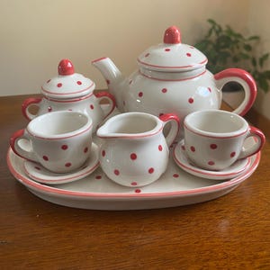 May include: A white ceramic tea set featuring a red polka dot pattern and red trim. The set includes a teapot with a lid, a sugar bowl with a lid, a creamer, two teacups with matching saucers, and an oval serving tray, all arranged together.