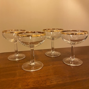 May include: A set of four clear glass coupe glasses, each featuring a wide, shallow bowl and a slender stem. The rim of every glass is finished with a thin, metallic gold band, providing a consistent decorative detail across the entire collection.