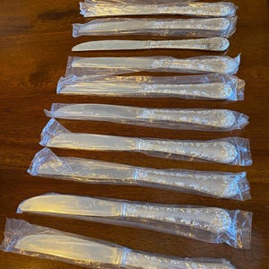 May include: A collection of twelve silver-toned dinner knives, each individually wrapped in clear plastic packaging. The knives feature a smooth, polished blade and a decorative, textured handle with an embossed floral pattern along the edges.