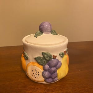 May include: A white ceramic canister with a matching lid topped by a purple plum handle. The exterior features a raised, hand-painted design of various fruits including orange slices, a cluster of purple grapes, and a yellow pear with green leaves.