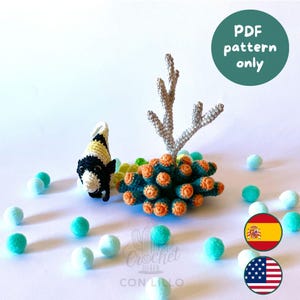 May include: A crochet pattern PDF featuring a black, white, and yellow fish next to a grey branching coral structure and a cluster of dark green and orange coral polyps. Small light blue and teal pom-poms are scattered around the base.