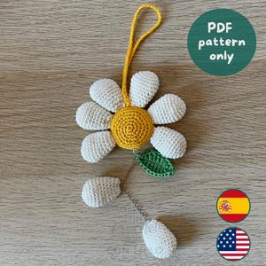 Puede incluir: Un adorno de margarita tejido a ganchillo con pétalos blancos, un centro amarillo y una hoja verde. Un lazo de hilo amarillo sobresale por la parte superior y una cadena plateada cuelga de la parte inferior, uniendo dos pequeñas esferas blancas tejidas. Texto: PDF pattern only.