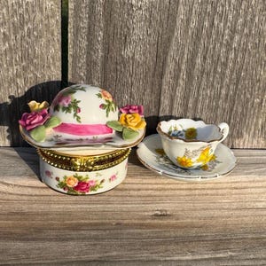 Puede incluir: Una caja de baratijas decorativa de porcelana con forma de sombrero con una cinta rosa y detalles florales, junto con una taza y un platillo a juego. La caja del sombrero es blanca con detalles florales rosas, amarillos y verdes.