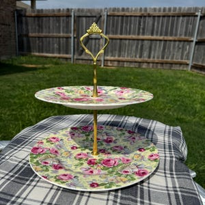 May include: Two-tiered serving tray with a gold-colored handle and stem. The plates feature a floral pattern of pink roses on a pale yellow background. The tray is set on a black and white checkered cloth, outdoors.