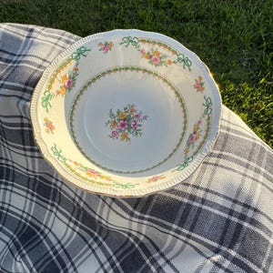 May include: A round ceramic bowl with a scalloped edge, featuring a white base, a central floral bouquet in pink and yellow, and a decorative border of green ribbons and small flowers along the interior rim with a gold-toned trim.