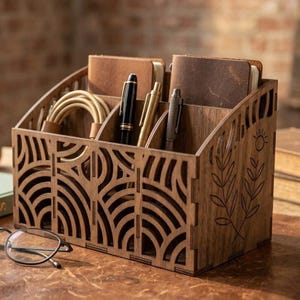 May include: A wooden desk tidy with several compartments. The light brown wood organiser has decorative cut-out patterns on the front. It contains pens, notebooks, and a charging cable. A sun and leaf design is etched on the side.