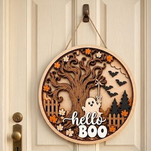 May include: A circular wooden wall hanging featuring a layered design of a large tree, a ghost holding a rope, bats, pine trees, and a fence. The text "hello BOO" is displayed in white script and block lettering. Orange maple leaves accent the scene.