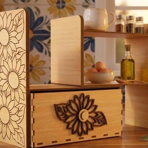 May include: A light-colored wooden desktop organizer featuring a drawer with a dark brown sunflower cutout and a side panel engraved with three sunflowers. The unit includes an upper shelf holding a ceramic mug, three spice jars, and a bowl of eggs.