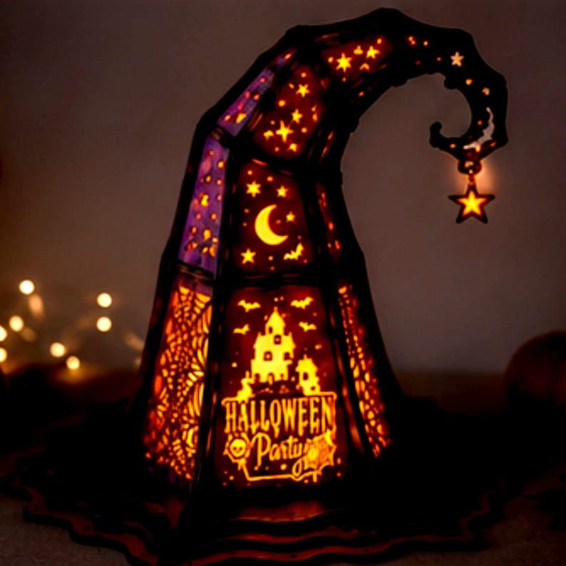 May include: A conical lamp shaped like a witch hat with a dark frame and glowing orange panels. The panels feature cutouts of a crescent moon, stars, bats, a haunted house, and spiderwebs. The text HALLOWEEN PARTY is visible, and a star charm hangs from the tip.