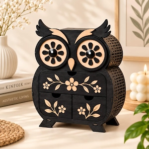 May include: A black wooden owl-shaped storage organizer featuring two drawers and a top compartment. The owl has large circular eyes with flower-shaped pupils, a pointed beak, and light-colored floral vine cutouts decorating the drawer fronts.