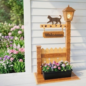 May include: A wooden cat-themed welcome sign with a picket fence and a planter box filled with colorful flowers. The sign features a cat silhouette, paw prints, and the word "Welcome". A decorative lamp is on top.