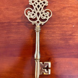 May include: A decorative brass key with an ornate, openwork design at the top. The key has a long, slender stem and a detailed key bit. The key is a warm gold color and is set against a reddish-brown background.