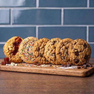 May include: A row of six round cookies on a wooden board. The cookies include a jam-topped cookie, a colorful sprinkle cookie, two oatmeal raisin cookies, and two chocolate chip cookies, with scattered ingredients like chocolate chunks.