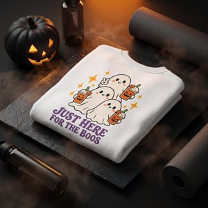 Peut inclure: Un sweat-shirt blanc à col rond orné d'un motif représentant trois fantômes de dessin animé tenant des boissons sur le thème de la citrouille. Le texte "JUST HERE FOR THE BOOS" est imprimé en violet sous les fantômes, avec des étoiles jaunes.