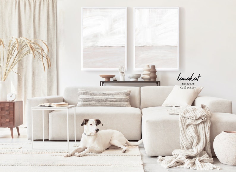 May include: A white sectional sofa with a white throw blanket and a white side table. A white dog is lying on a cream-coloured rug in front of the sofa. Two abstract paintings in white and beige frames are hanging on the wall above the sofa. The text "lanakat Abstract Collection" is printed on the wall.