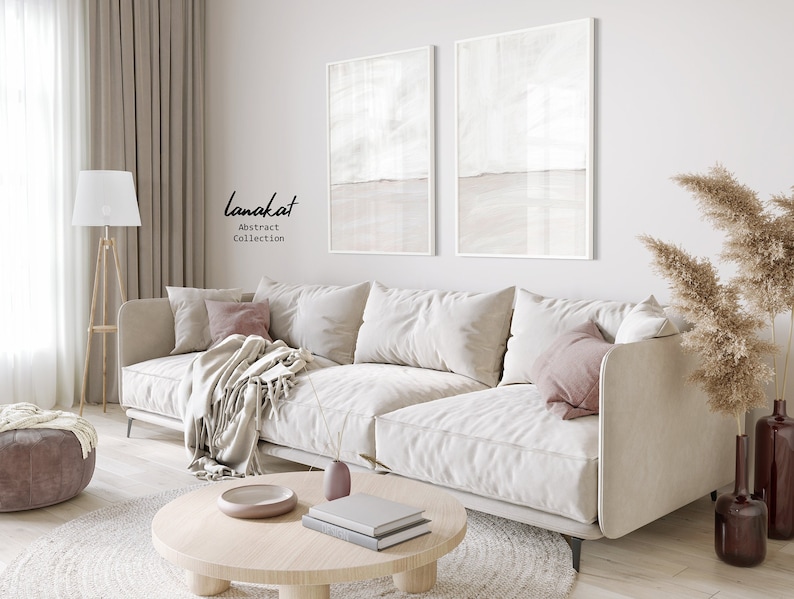 May include: A white sectional sofa with pink throw pillows and a white throw blanket sits in a living room with a wooden coffee table, a floor lamp, and a vase of dried pampas grass. The wall behind the sofa features two abstract paintings with a white and beige colour scheme. The text "lanakat Abstract Collection" is visible on the wall.