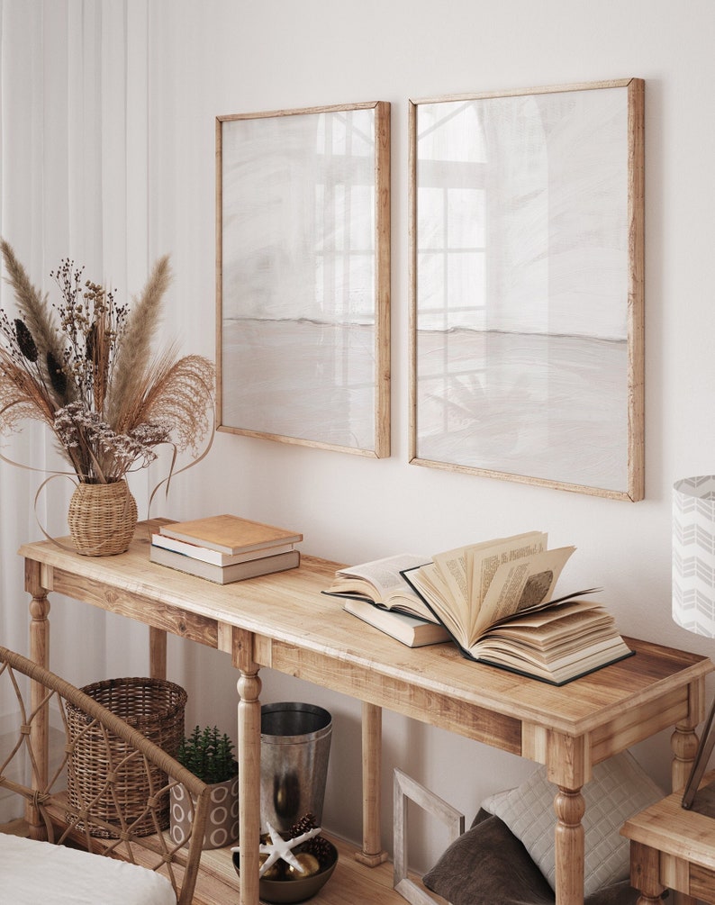 May include: A wooden console table with a light wood finish. The table has two shelves and is decorated with a vase of dried flowers, books, and a decorative bowl. Two framed prints with a light beige background hang on the wall behind the table.