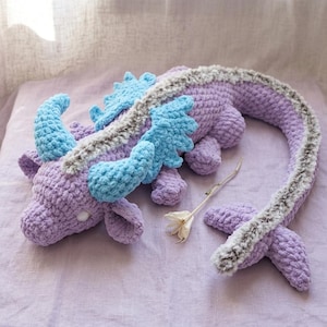 Può includere: Un drago all'uncinetto fatto a mano nei toni del lavanda, azzurro e grigio. Il peluche presenta una lunga coda, corna e ali. Il drago è adagiato su una superficie lavanda, con un fiore secco nelle vicinanze.