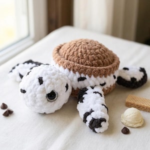 May include: Crocheted ice cream sandwich toy with white and brown yarn. The toy has a round shape with a brown top and bottom, white filling, and black spots. Additional crocheted elements include a scoop of ice cream, chocolate chips, and a wafer.