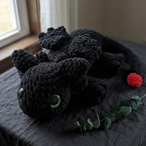 May include: A handmade black crochet dragon plushie with large green eyes and a red pom-pom tail. The dragon is lying on a dark gray surface, with a sprig of greenery nearby. The plushie is a soft, cuddly toy.