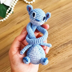 May include: A handmade blue crochet toy shaped like a cartoon character with large ears and a long, twisted body. The toy is light blue with darker blue accents on the ears, feet, and nose. The eyes are black and white.