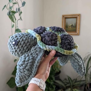 May include: A handcrafted crochet turtle in grey, blue, and green hues. The plush turtle features a textured shell and flippers, with a white eye detail. A soft, cuddly toy.