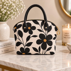 Könnte beinhalten: Eine kuppelförmige Handtasche mit cremefarbener Basis, einem auffälligen schwarzen Blumenmuster und kleinen gelben Blütenmitten. Die Tasche verfügt über zwei schwarze Tragegriffe, einen goldfarbenen Reißverschluss mit rundem Zipper und goldene Beschläge.