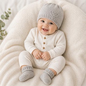 Peut inclure: Un bébé souriant portant un bonnet en tricot gris clair avec un motif de cœur, un body crème avec des boutons en bois et des chaussettes en tricot gris assorties. Le bébé est assis sur une couverture en tricot crème.
