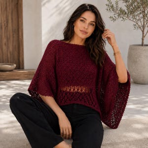 May include: A deep burgundy, loose-fitting knit sweater featuring an intricate openwork crochet pattern, a wide boat neckline, and flowing, elbow-length sleeves. The garment is paired with solid black trousers, creating a contrast in texture and color.