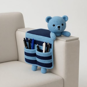 May include: A blue and navy blue crocheted organizer with two pockets, shaped like a bear, holding pens. The bear is perched on the arm of a light-colored sofa. The organizer has a striped pattern.