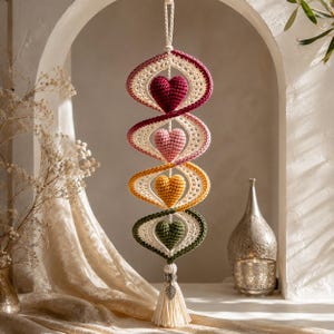 May include: A vertical hanging decoration featuring four crocheted hearts in dark red, light pink, yellow, and dark green. Each heart is encased in a spiraling, cream-colored crocheted frame. The piece ends with a cream tassel and a silver leaf charm.