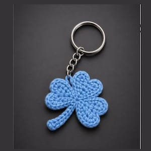 May include: A light blue crocheted four-leaf clover keychain with a silver-colored keyring and chain. The clover is made of a textured yarn and is set against a dark background.