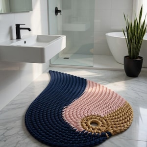 Peut inclure: Tapis de bain en forme de goutte d'eau, composé de fils épais tissés en bleu marine, rose blush et beige. Le tapis est posé sur un sol carrelé en marbre dans une salle de bain moderne.
