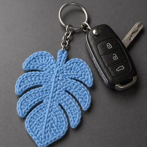 May include: A light blue, crocheted leaf keychain with a silver keyring and chain. A black car key fob and car key are next to the keychain. The leaf design is a tropical monstera leaf.