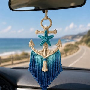 May include: A macrame car charm shaped like an anchor, crafted from cream-colored cord with a teal starfish accent at the center. The bottom of the anchor features a long, layered fringe that transitions in color from light blue to deep navy blue.