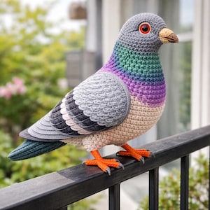 May include: A handmade crochet pigeon with a gray head, wings, and tail, and a multicolored chest. The bird has orange feet and a tan beak, perched on a black railing. The background is blurred with greenery.