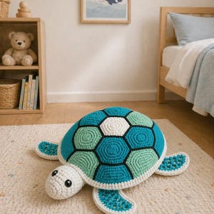 Pode incluir: Uma almofada de tartaruga de crochê feita à mão em tons de azul, verde e branco. A tartaruga tem uma carapaça estampada, cabeça branca e barbatanas azuis. É colocada sobre um tapete creme em um quarto infantil.