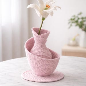 May include: A handmade crochet vase set in a soft pink hue, featuring a unique spiral design. The set includes a vase, a bowl, and a coaster, with a white crochet lily flower. The set is displayed on a marble table.