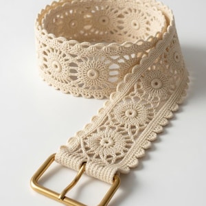 May include: Cream-coloured crochet belt with a gold-coloured rectangular buckle. The belt features a floral pattern and a scalloped edge. The belt is rolled up on a white background.
