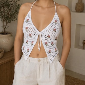 Op de afbeelding: Witte gehaakte haltertop met roze bloemenborduurwerk. De top heeft een striksluiting bij de buste en een delicaat, open weefselontwerp. Gecombineerd met lichtgekleurde linnen broek, wat een casual, zomerse uitstraling creëert.