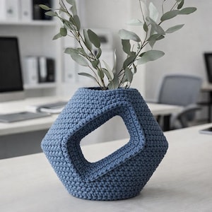 Op de afbeelding: Een blauwe gehaakte vaas met een geometrisch ontwerp, met een grote rechthoekige opening. De vaas bevat groene, bladrijke takken. De vaas staat op een wit oppervlak, met kantoormeubilair op de achtergrond.