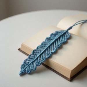 May include: A blue crochet feather bookmark rests on the pages of an open book. The bookmark has a detailed, textured design with a scalloped edge. The book has a brown cover and cream-colored pages. The bookmark is approximately 8 inches long.