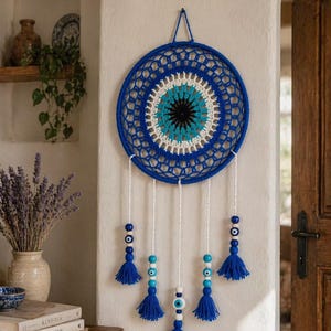 May include: A blue and white crochet wall hanging with a central eye design. The circular piece has a blue outer ring and a detailed inner pattern in white, turquoise, and black. White strings with blue tassels and beads hang below, creating a decorative piece.
